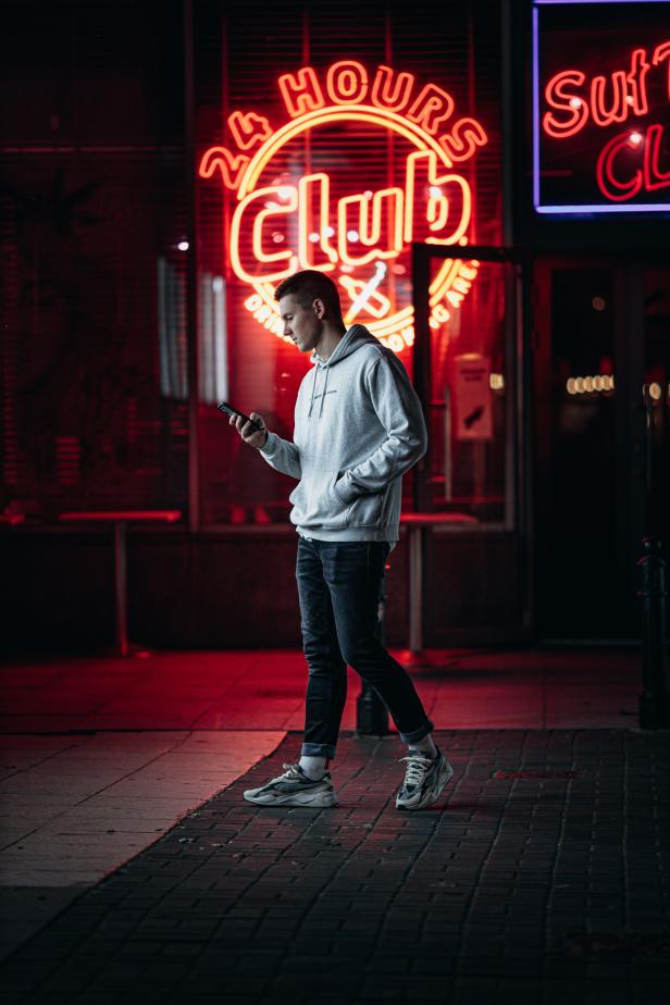 Ein junger Mann geht nachts an einem „Club“-Schild vorbei und schaut auf sein Handy.