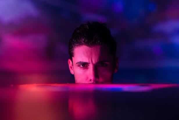 Ein Mann, bis zur Nase im Wasser, blickt ernst in die Kamera; violettes Licht im Hintergrund.