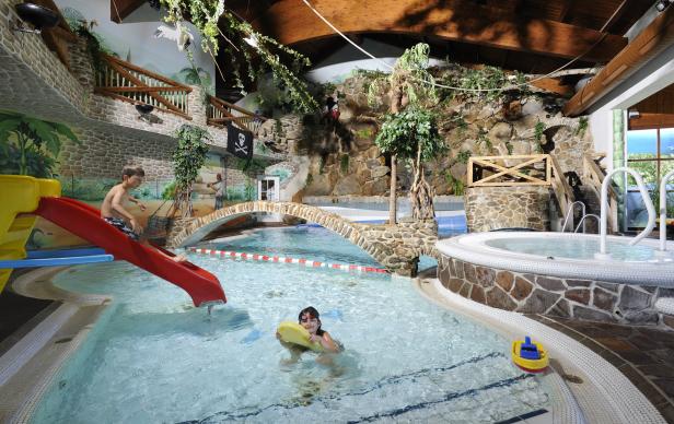 Ein Junge rutscht eine rote Rutsche hinunter, während ein Mädchen mit einem Schwimmbrett im Wasser schwimmt.