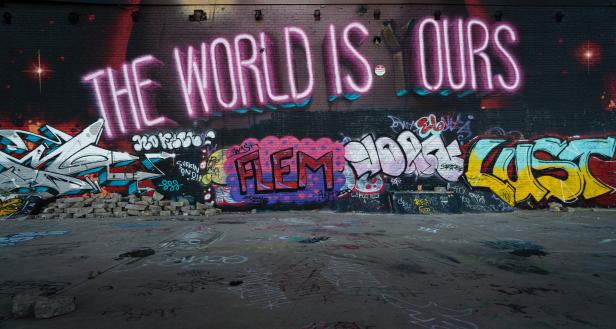 Eine mit Graffiti bedeckte Wand mit der Leuchtschrift „The World is Ours“.