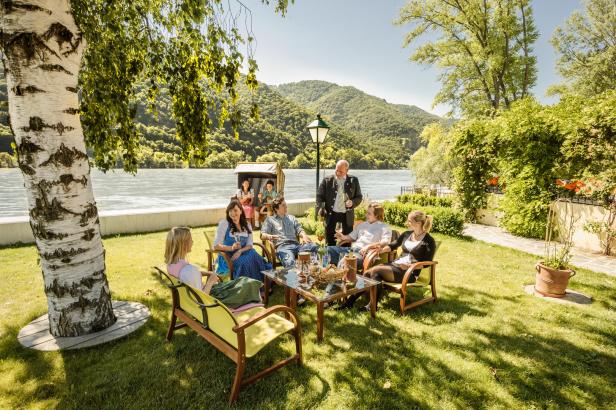 Eine Gruppe von Personen sitzt im Freien und genießt Wein mit Blick auf einen Fluss und bewaldete Hügel.