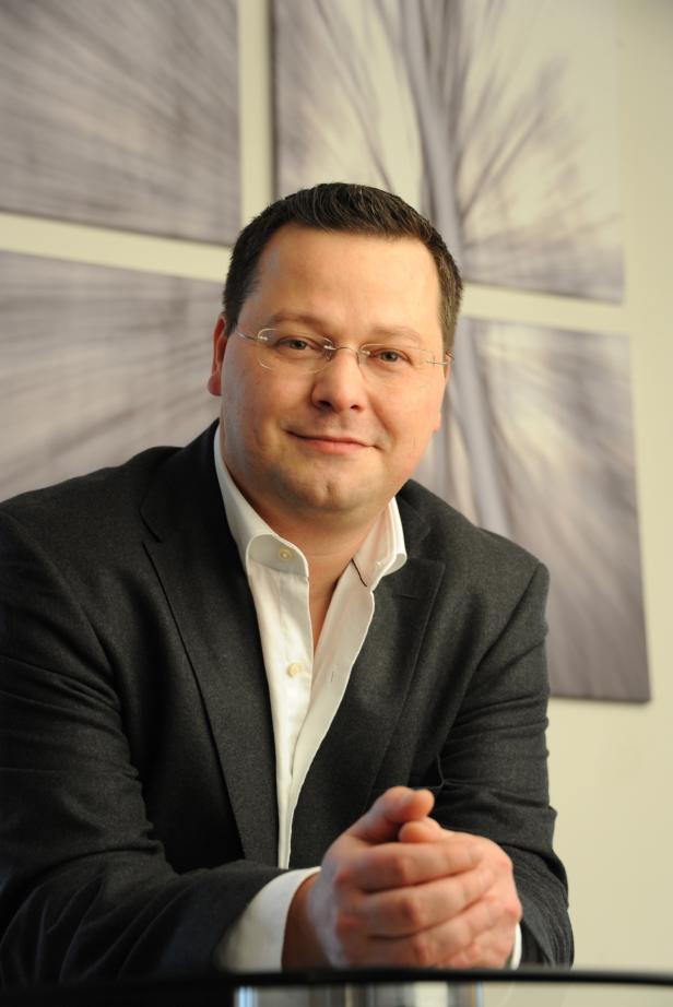 Ein Mann mit Brille und dunklem Anzug sitzt vor einem abstrakten Hintergrund.