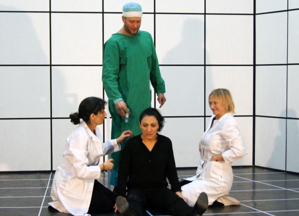 Eine Frau sitzt auf dem Boden, während zwei Ärztinnen und ein Arzt in grüner OP-Kleidung um sie herum stehen.