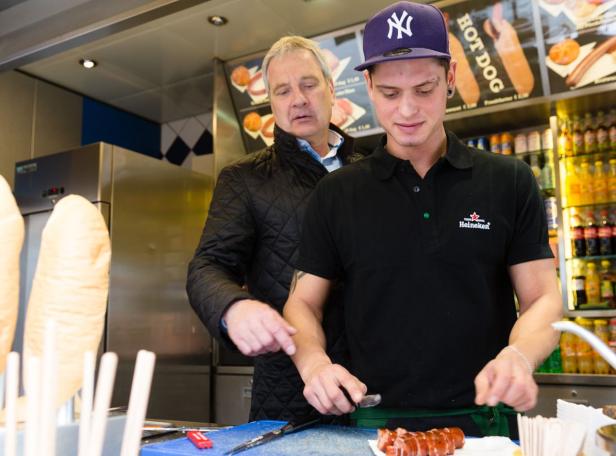 Ein junger Mann mit New-York-Yankees-Kappe schneidet Würstchen an einem Imbissstand.