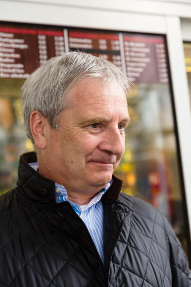 Ein Mann mit grauen Haaren lächelt vor einem Fenster mit einer Speisekarte.