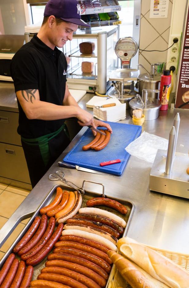 Ein Mann schneidet Würstchen auf einem blauen Schneidebrett in einer Imbissküche.