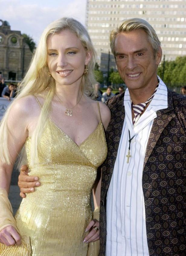Eine blonde Frau in einem goldenen Kleid posiert mit einem Mann vor einem Gebäude.