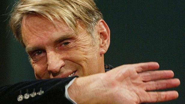 Ein Mann mit blonden Haaren lächelt und hält seine Hand vor das Gesicht.