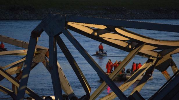 Mehrere Personen in roten Overalls fahren mit Schlauchbooten auf einem Fluss, gesehen durch eine Stahlbrückenkonstruktion.