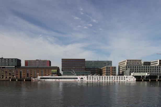 Ein Flusskreuzfahrtschiff liegt vor modernen Bürogebäuden vor Anker.