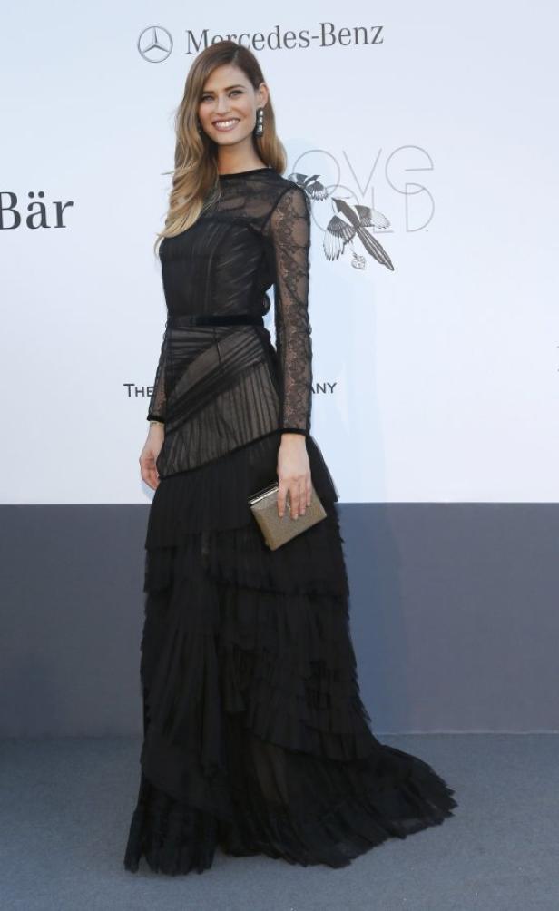 Eine Frau in einem langen, schwarzen Kleid mit Spitze und Rüschen hält eine goldene Clutch.