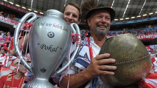 Zwei Bayern-Fans mit einem aufblasbaren Pokal und einer Kuhglocke im Stadion.
