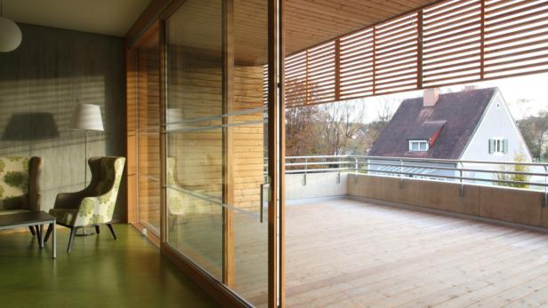 Blick von einem Zimmer mit Sessel auf eine Holzterrasse mit einem Haus im Hintergrund.