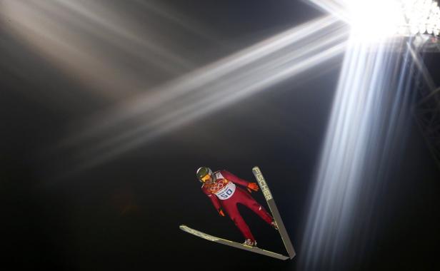 Ein Skispringer in rotem Anzug springt vor hellem Scheinwerferlicht.