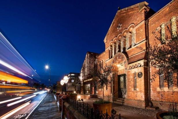 Das Manchester Jewish Museum in einem Backsteingebäude bei Nacht mit verwischten Lichtern vorbeifahrender Autos.