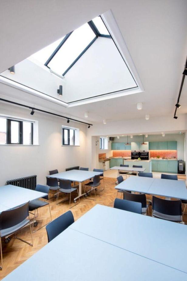 Ein heller Raum mit Tischen und Stühlen, im Hintergrund eine Küche.