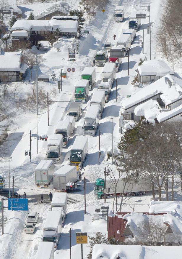 Schneebedeckte Straße mit einer Schlange von Lastwagen und Häusern in einer Winterlandschaft.