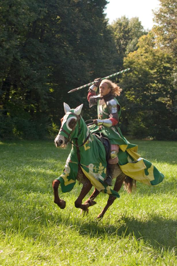 Ein Ritter in Rüstung reitet auf einem geschmückten Pferd und hält eine Lanze.