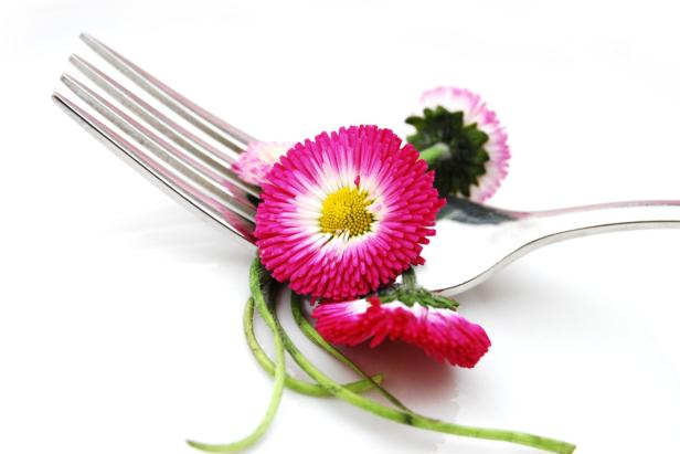 Eine Gabel liegt auf einem weißen Untergrund, dekoriert mit pinkfarbenen Gänseblümchen.
