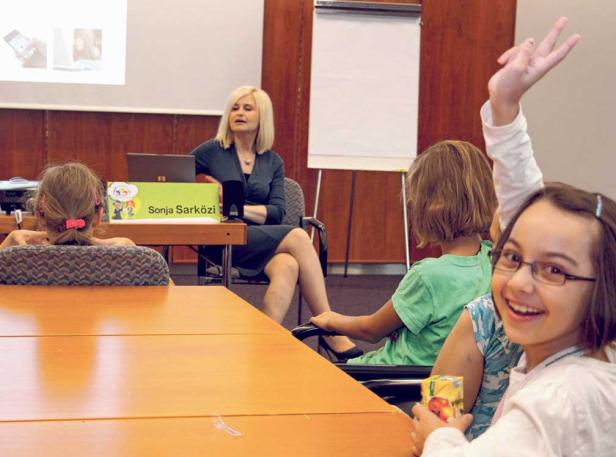 Sonja Sarközi hält einen Vortrag vor einer Gruppe von Kindern, von denen eines die Hand hebt.