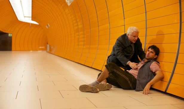 Ein Mann in Tracht liegt in einer U-Bahn-Station, während ein anderer sich um ihn kümmert.