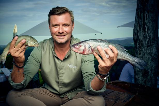 Christof Widakovich präsentiert zwei Fische vor einer malerischen Landschaft.
