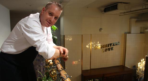 Ein Koch in Kochuniform lehnt an einem Geländer mit der Aufschrift „Schnitzel Love“ im Hintergrund.