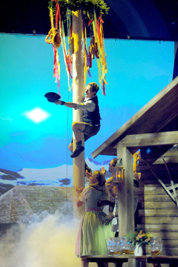 Ein Mann klettert auf einen Maibaum mit Brezeln und Würsten, während Zuschauer applaudieren.