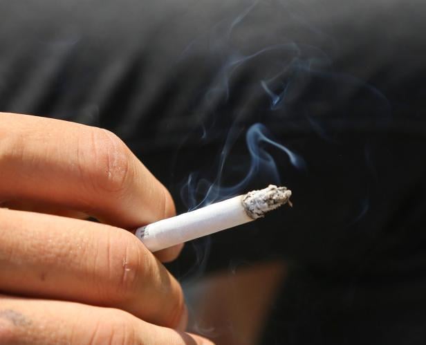 Eine Hand hält eine brennende Zigarette, von der Rauch aufsteigt.