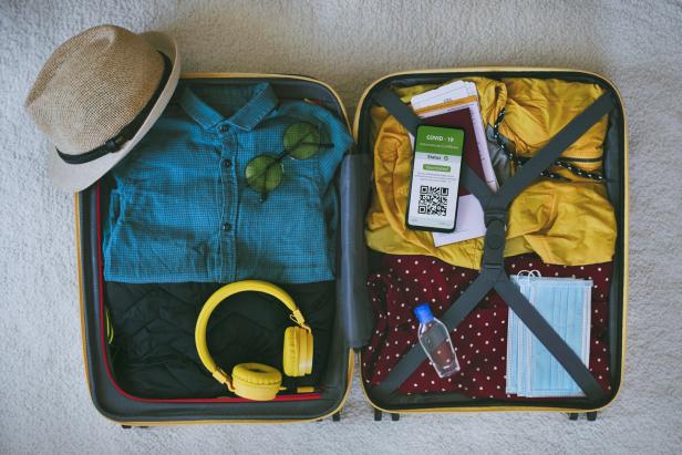 Ein offener Koffer mit Kleidung, einem Hut, Kopfhörern, einem COVID-19-Impfzertifikat und Masken für eine Reise.