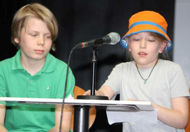 Zwei Kinder stehen an einem Tisch mit einem Mikrofon und lesen etwas vor.