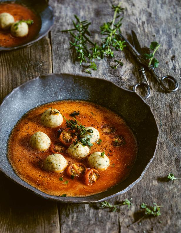 Eine Schüssel mit Tomatensuppe und Kräutern, garniert mit Knödeln und Tomatenstücken.