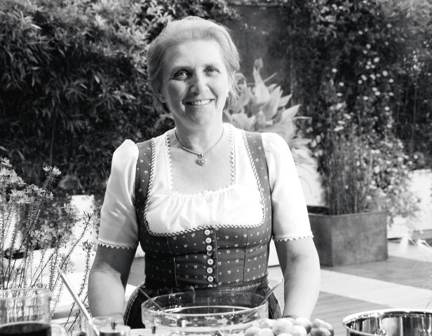 Eine lächelnde Frau im Dirndl steht vor einem Tisch mit Zutaten zum Kochen.