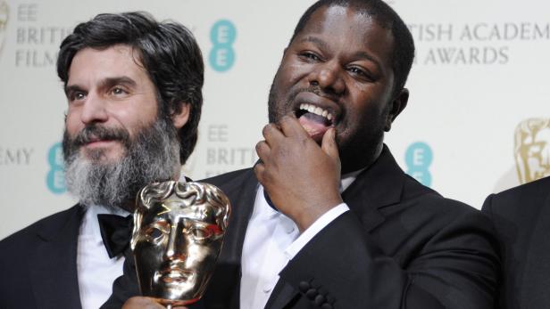 Steve McQueen hält eine BAFTA-Trophäe, während ein Mann mit Bart daneben steht.