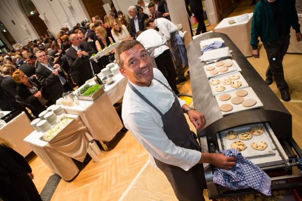 Ein lächelnder Koch steht vor einem Ofen mit Fladenbroten, im Hintergrund eine Menschenmenge.
