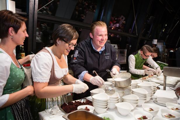 Ein Koch und mehrere Personen in Tracht bereiten Speisen in einer Küche zu.