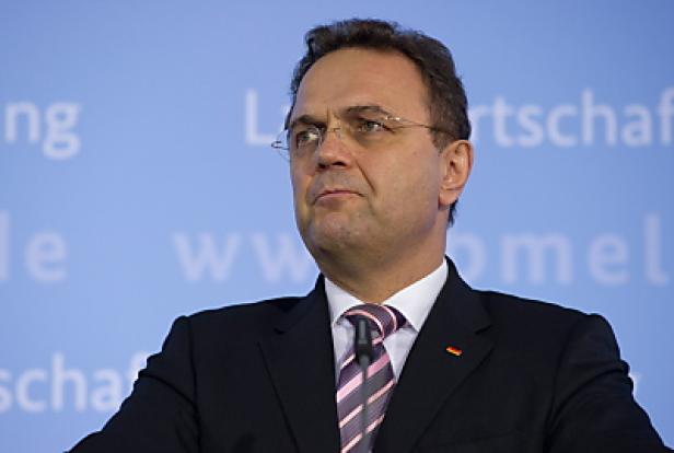 Ein Mann mit Brille und Anzug steht vor einem hellblauen Hintergrund.