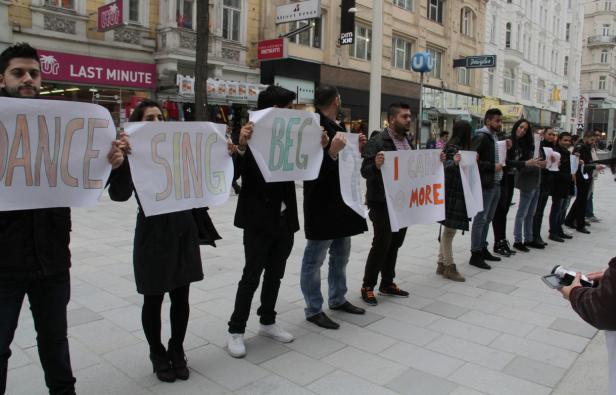 Eine Gruppe von Menschen hält Schilder mit den Aufschriften „Dance“, „Sing“, „Beg“ und „I can do more“.