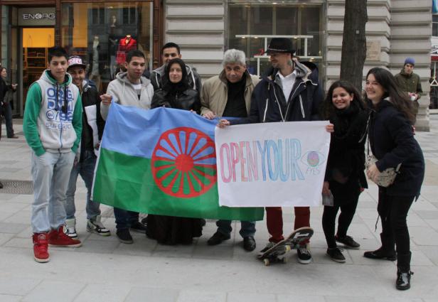 Eine Gruppe von Menschen hält eine Roma-Flagge und ein Schild mit der Aufschrift „Open Your Eyes“.