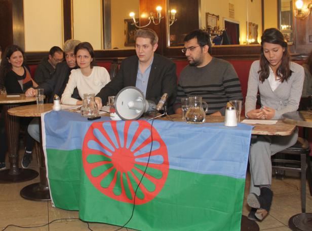 Eine Gruppe von Menschen sitzt an einem Tisch, der mit der Roma-Flagge bedeckt ist.