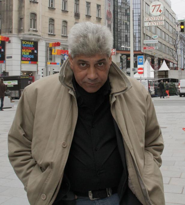 Ein Mann mit grauem Haar und beiger Jacke steht auf einem belebten Platz.