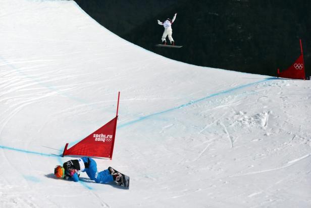 Eine Snowboarderin springt über eine Halfpipe, während eine andere gestürzt ist.