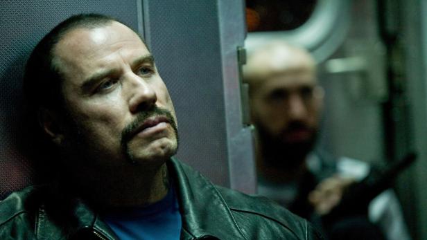 John Travolta in Lederjacke lehnt an einer Wand, während im Hintergrund eine weitere Person zu sehen ist.