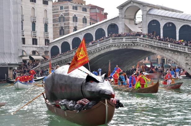 Boote mit Menschen in Kostümen fahren auf dem Canal Grande in Venedig unter der Rialtobrücke hindurch.