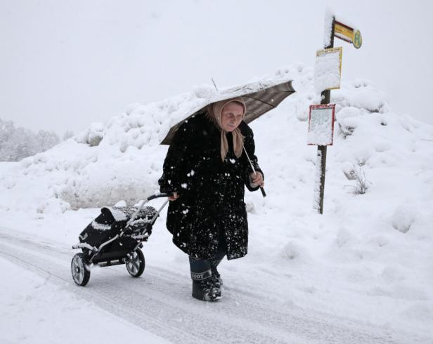 Eine ältere Frau geht mit einem Regenschirm und einem Kinderwagen durch eine verschneite Landschaft.