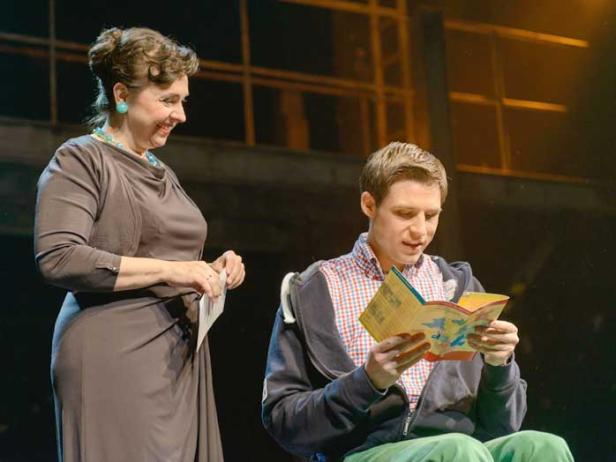 Eine Frau schaut lächelnd zu, wie ein junger Mann in einem Buch liest.