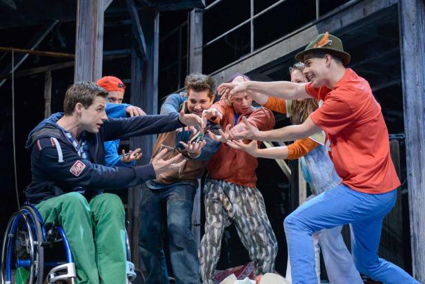 Eine Gruppe von Schauspielern auf einer Bühne, die um einen Schlüssel kämpfen; einer sitzt im Rollstuhl.