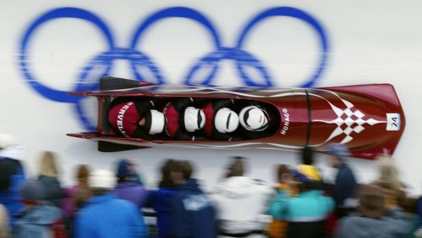 Ein Bobteam aus Monaco rast eine Eisbahn hinunter, im Hintergrund die olympischen Ringe.