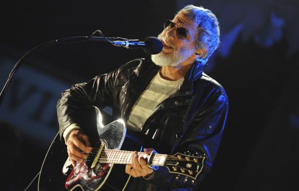 Ein älterer Mann mit Bart und Sonnenbrille spielt Gitarre und singt ins Mikrofon.
