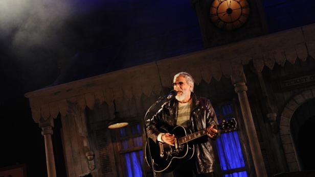 Ein Mann mit Gitarre singt vor einer Bahnhofskulisse.